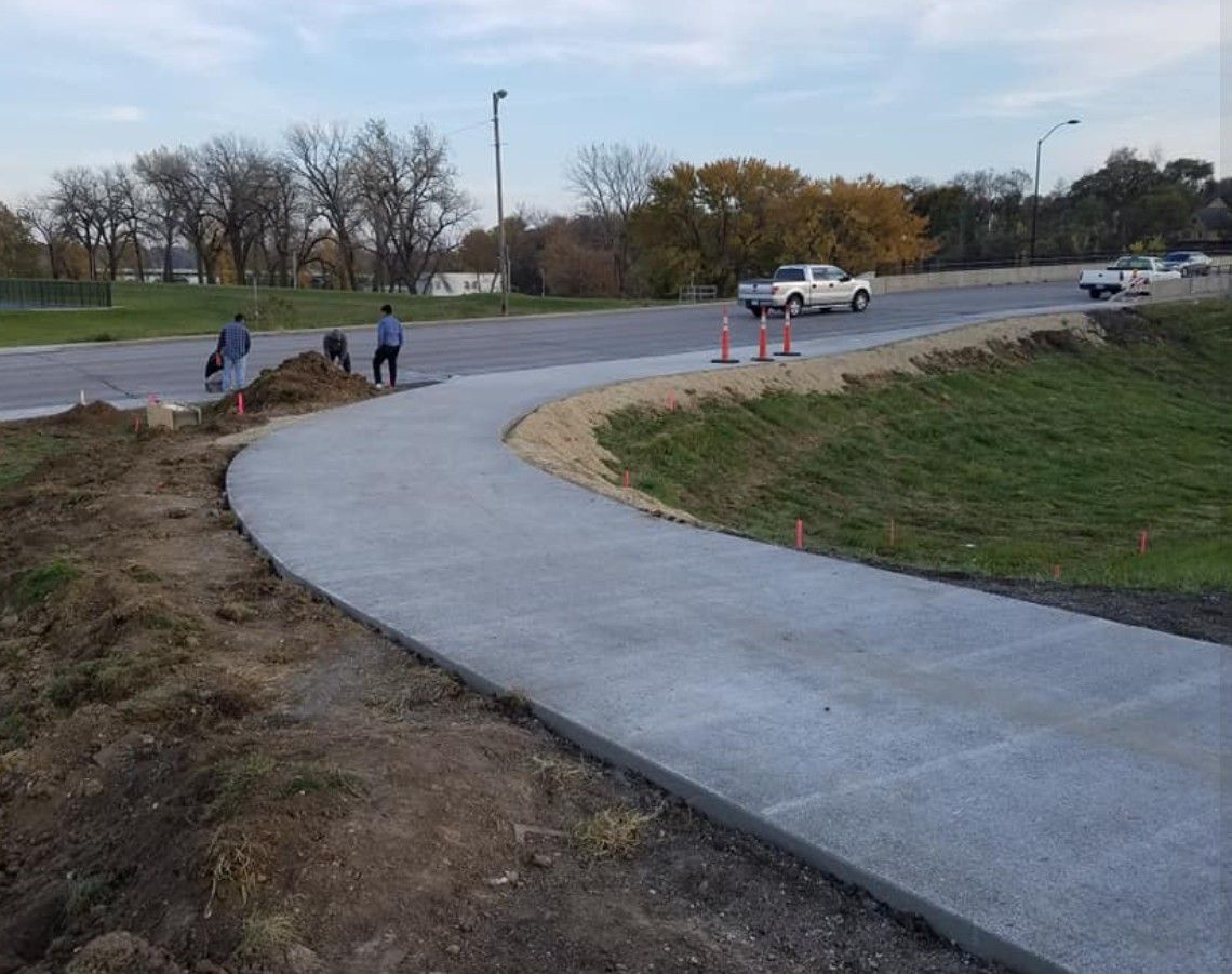 A newly paved concrete sidewalk curves along a grassy embankment near a road with construction workers and vehicles.