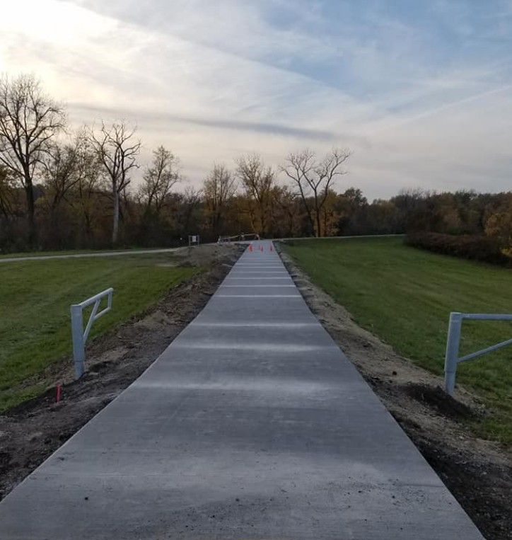A newly poured concrete path stretches into the distance, flanked by grassy fields, bare trees, and metal safety railings.