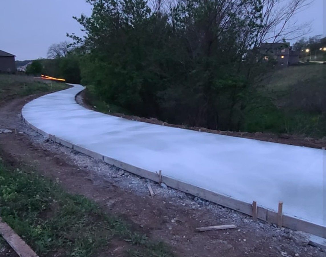 A newly poured concrete walking path curves through a grassy area at dusk, framed by wooden construction forms.