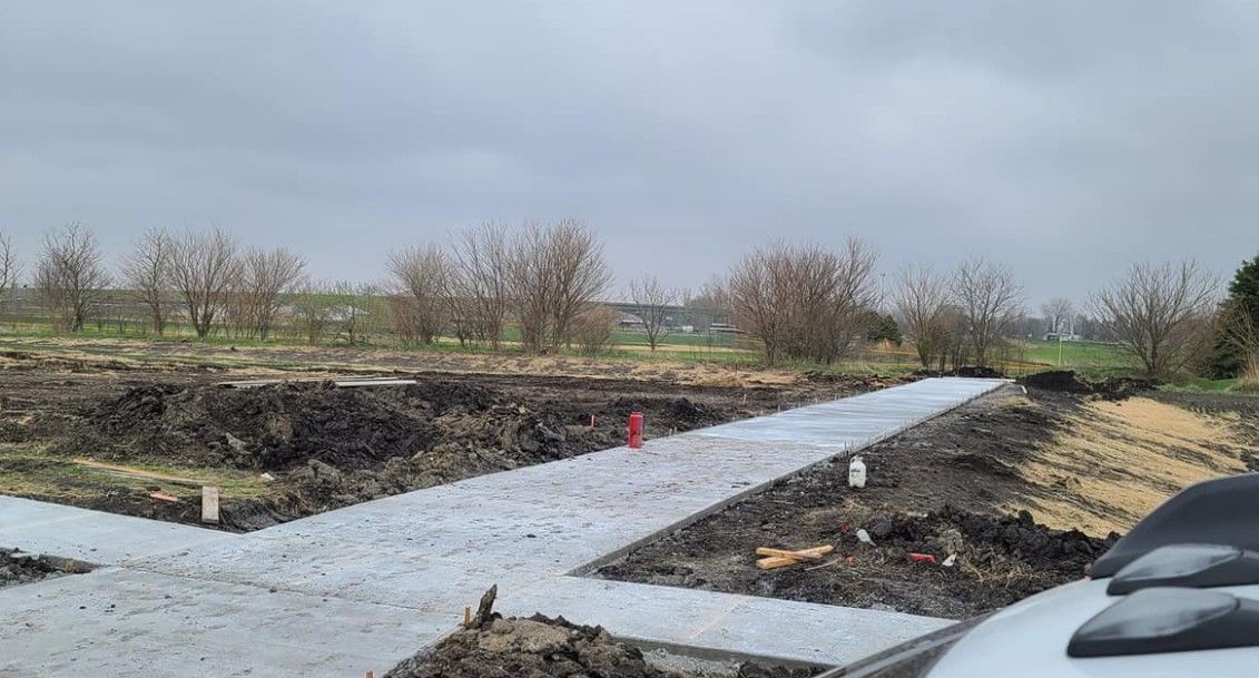 A newly poured concrete sidewalk intersecting across a muddy, undeveloped construction site under an overcast sky.