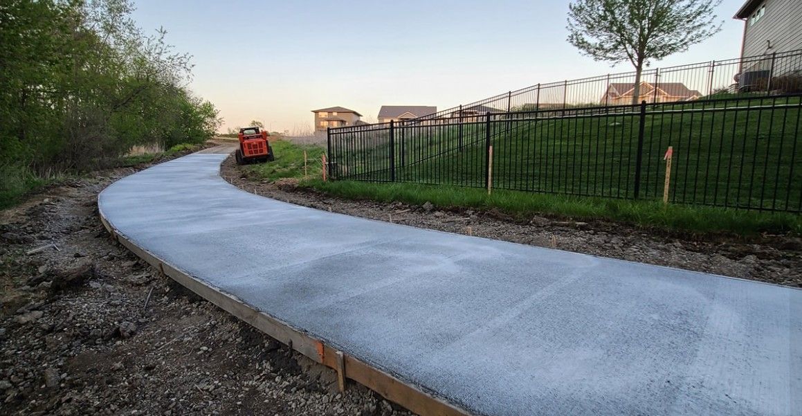 A freshly poured concrete sidewalk curves through a residential yard, bordered by a black fence and small construction stakes.