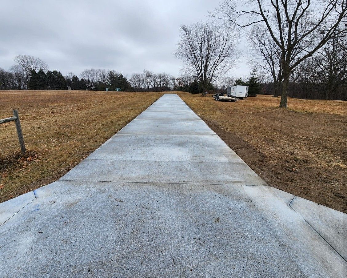 A long, straight concrete driveway stretches through a flat, brown grassy field under a cloudy sky.