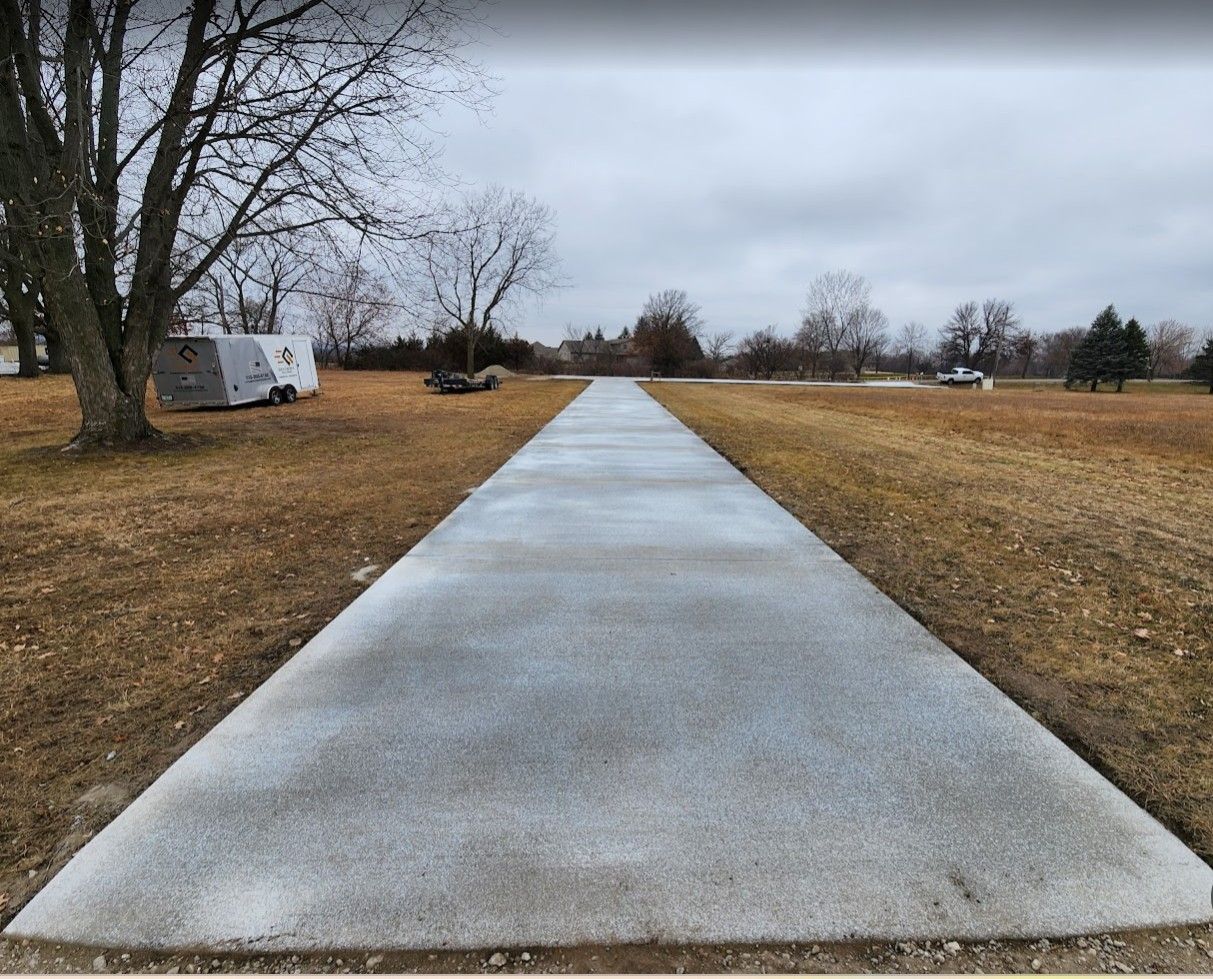 A newly poured, rectangular concrete path stretches through a flat, brown grassy field under a cloudy, overcast sky.