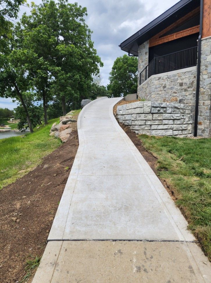 A paved sidewalk curves upward alongside a stone retaining wall and building, with trees and a grassy hillside nearby.