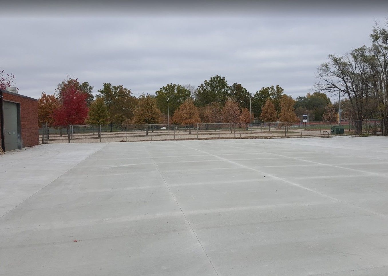 Empty concrete tennis courts with a fence and colorful trees in the background under a cloudy sky.