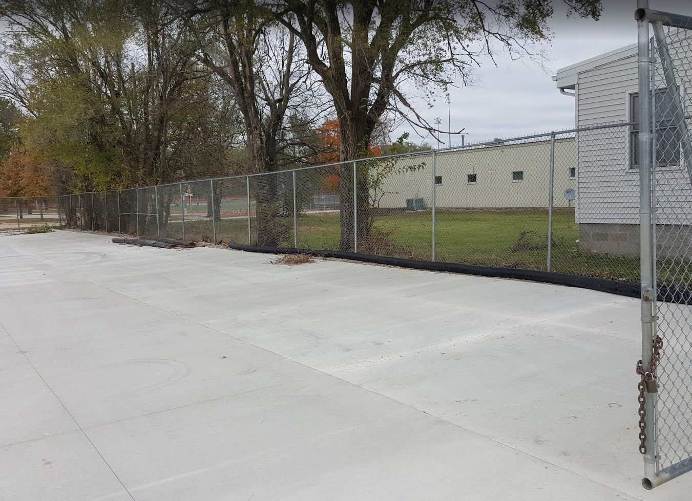 Concrete area next to a chain link fence and a building. Trees and grass visible. Overcast day.