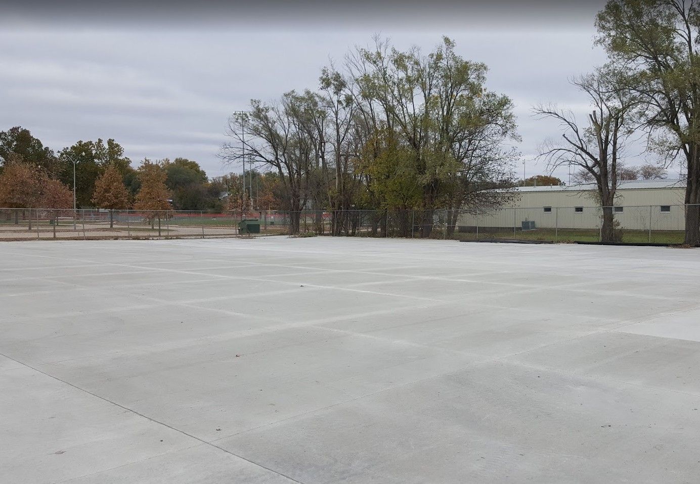 Empty concrete lot with trees and a building in the background under a cloudy sky.