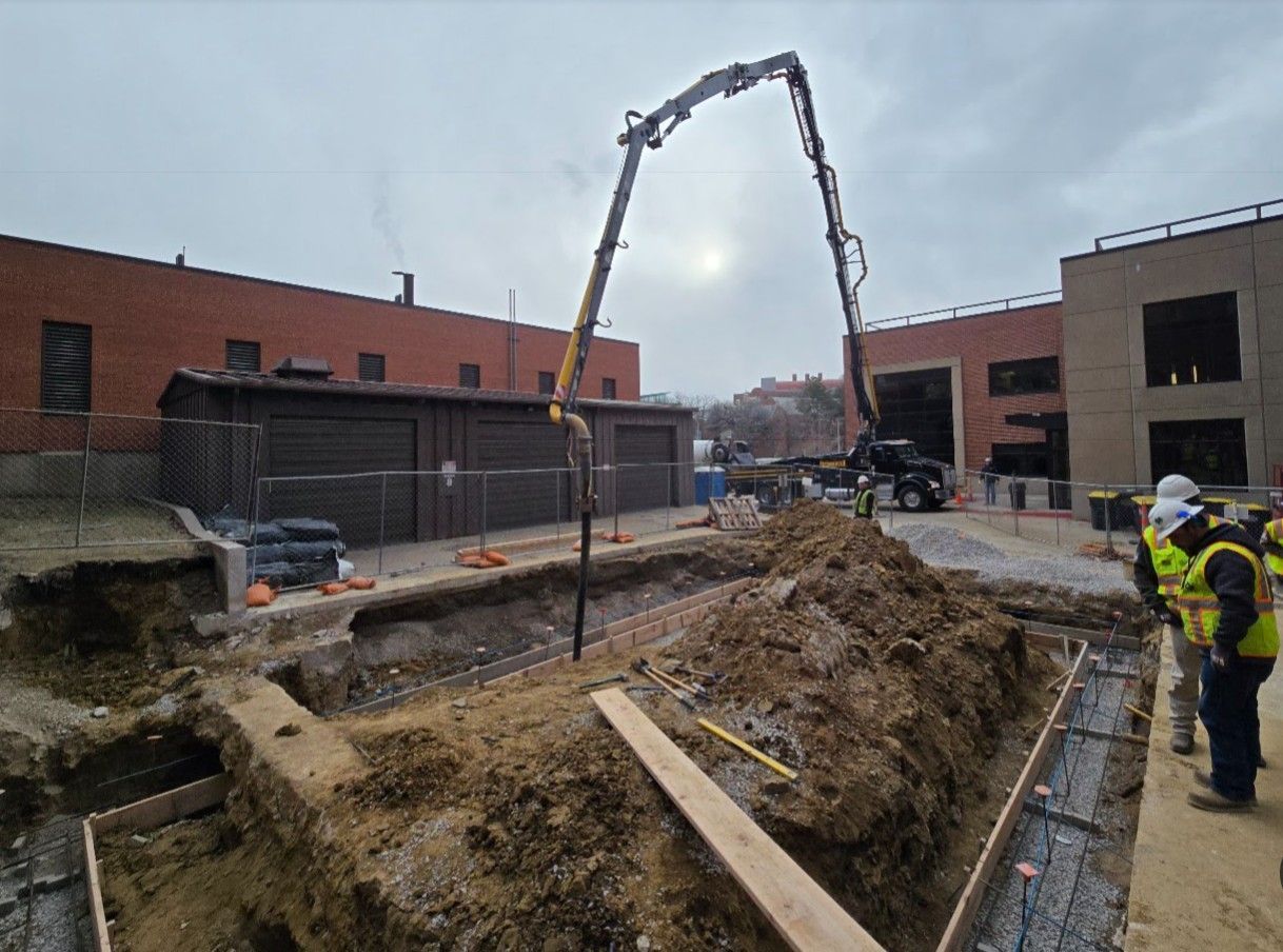 A construction crew operates a concrete pump truck at a job site with an excavated foundation and reinforced steel.