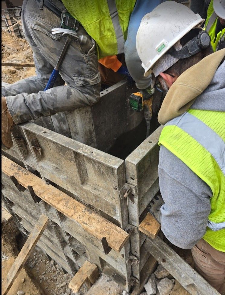 Construction workers in hard hats and high-visibility vests work on assembling metal concrete forms at a job site.