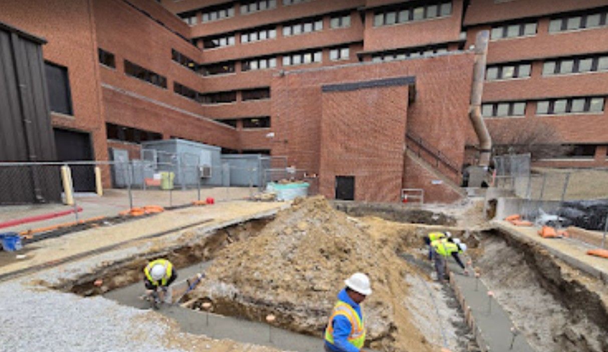 Construction workers level concrete in a trench at a brick building site with a large pile of excavated dirt.