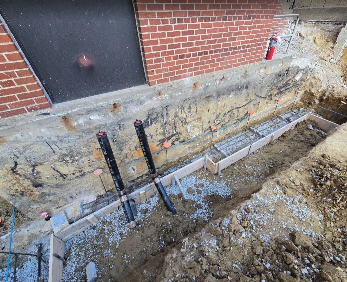 A construction trench featuring wooden concrete forms, metal rebar, and two vertical pipes along a brick foundation wall.