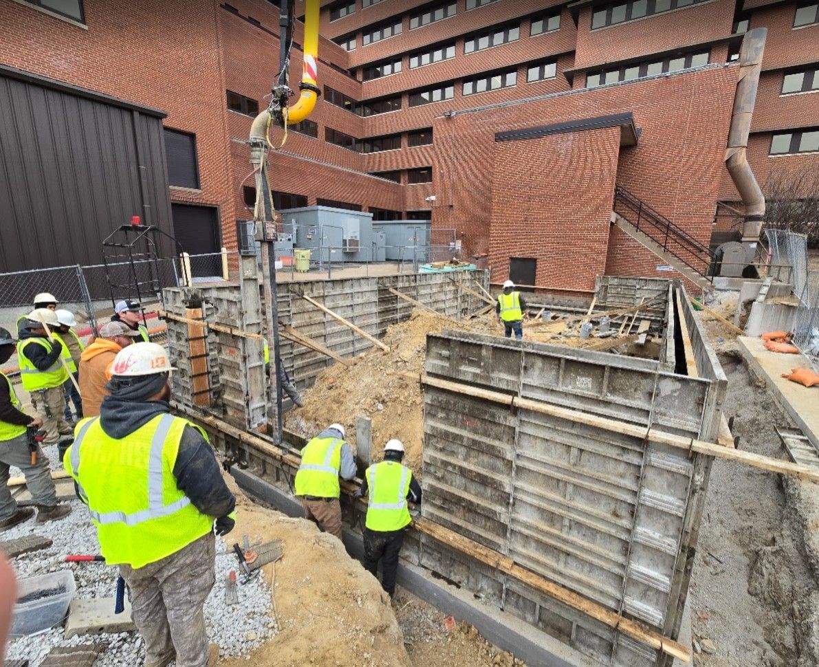Construction workers in high-visibility vests work at a building site with concrete forms and a pump hose.