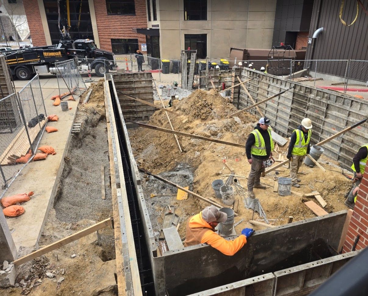 Construction workers in high-visibility vests work inside a foundation formwork at a building site.