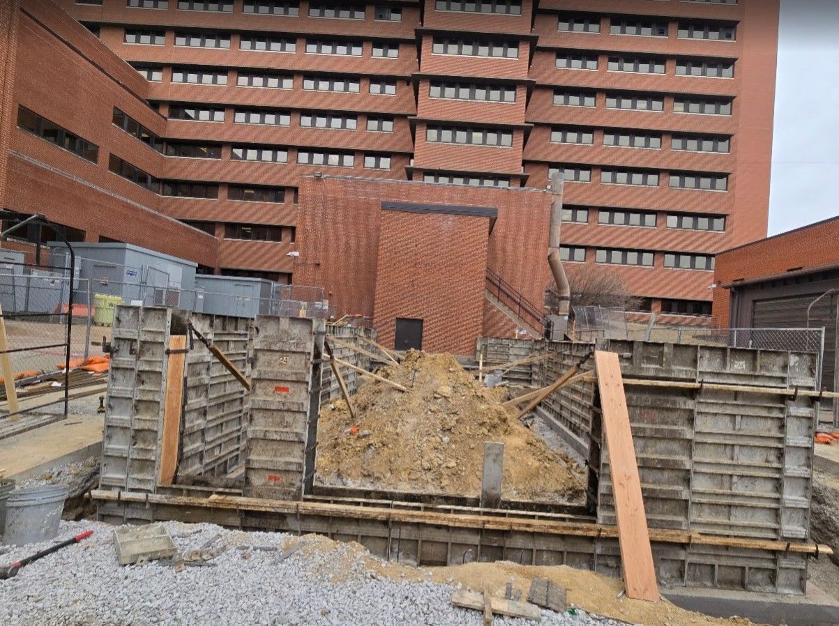 Construction site with metal concrete forms arranged in a rectangular trench filled with dirt in front of a brick building.
