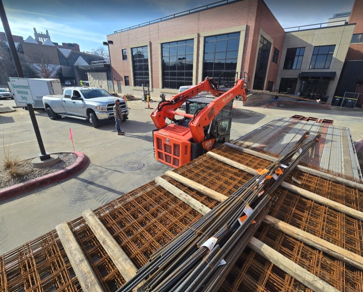 Construction site with a bright orange skid steer, a white pickup truck, and a flatbed trailer loaded with rebar.