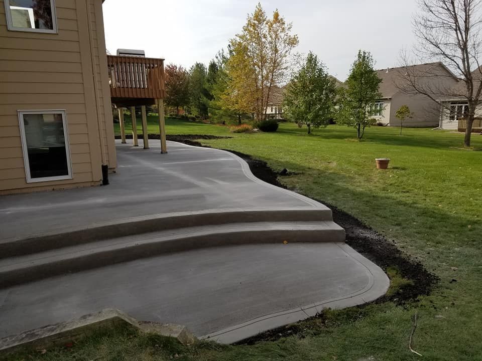 Concrete patio with two steps, adjacent to a house, edged with a dark soil bed, grassy yard.
