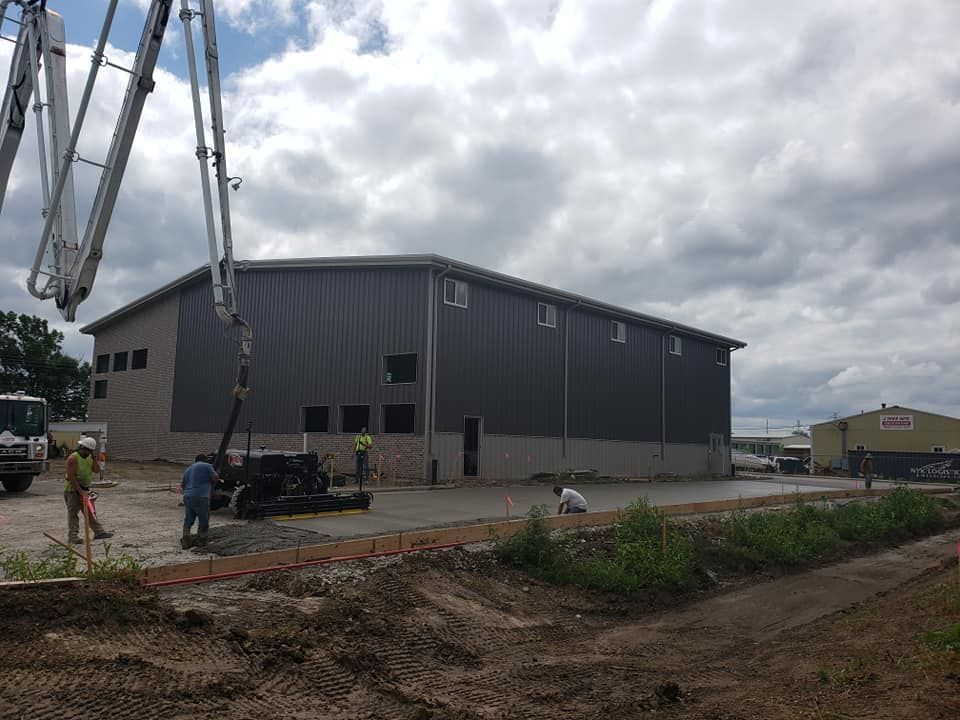Construction site with a concrete pump pouring concrete foundation for a gray metal building. Workers are present.