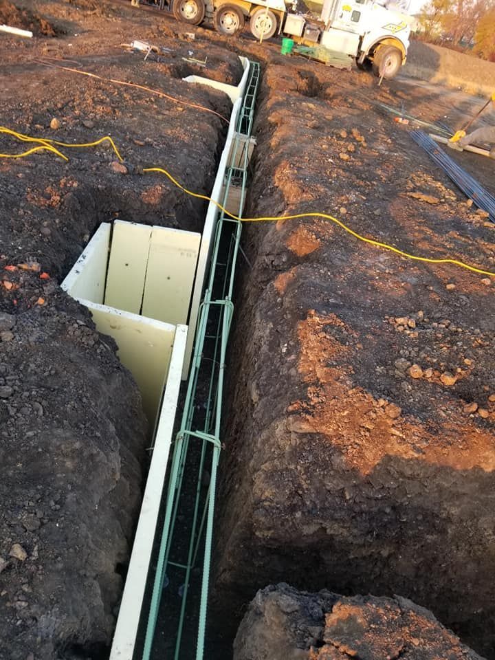 Construction site: Trench with metal reinforcement, green bars, and white foam walls; machinery in background.