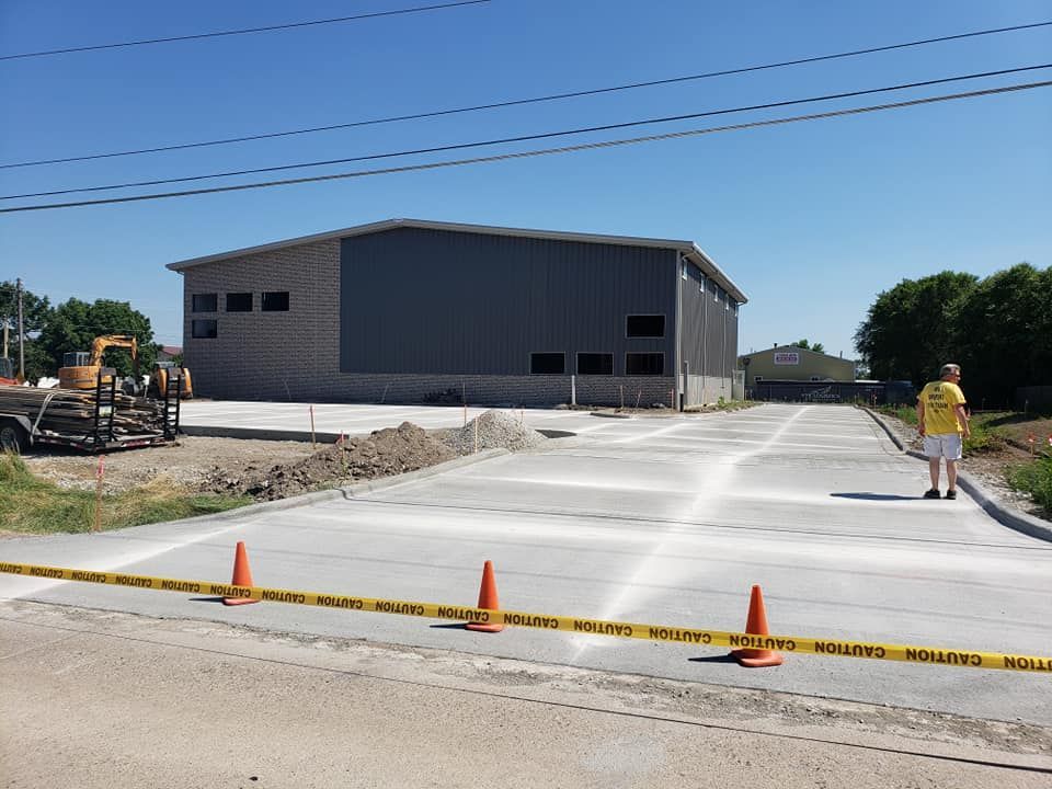 Construction site with a large gray building and newly poured concrete parking area. Orange cones and caution tape block access.