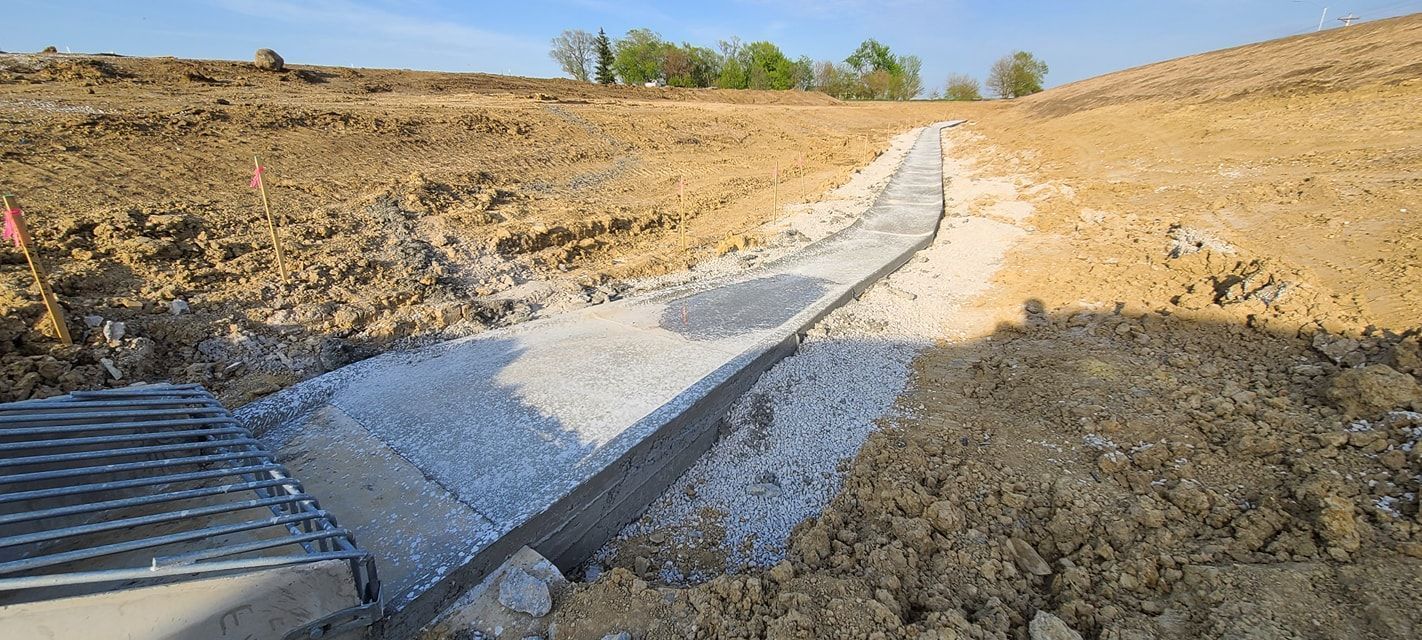 Construction site: drainage ditch with concrete and gravel, surrounded by brown earth.