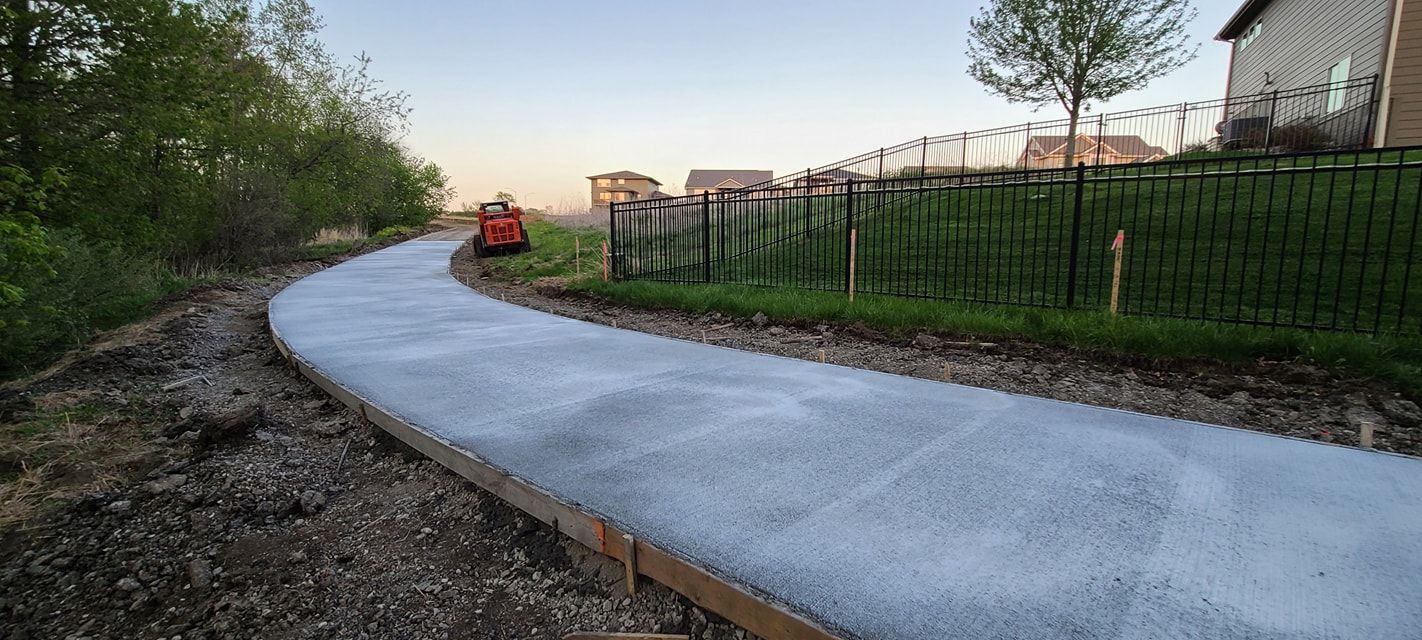 A concrete path curves through a grassy area, with a fence and trees to the right and left.