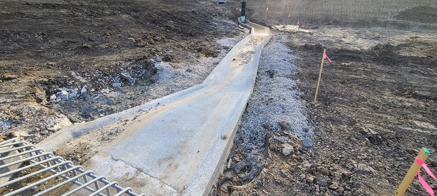 Concrete drainage channel in dirt, with a metal grate at the end. Pink tape on a stake to the right.