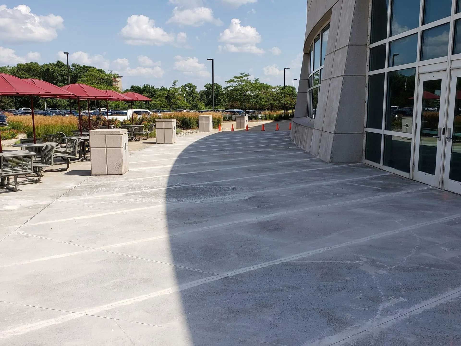 Outdoor patio with concrete walkway, tables, red umbrellas, and a modern building on a sunny day.