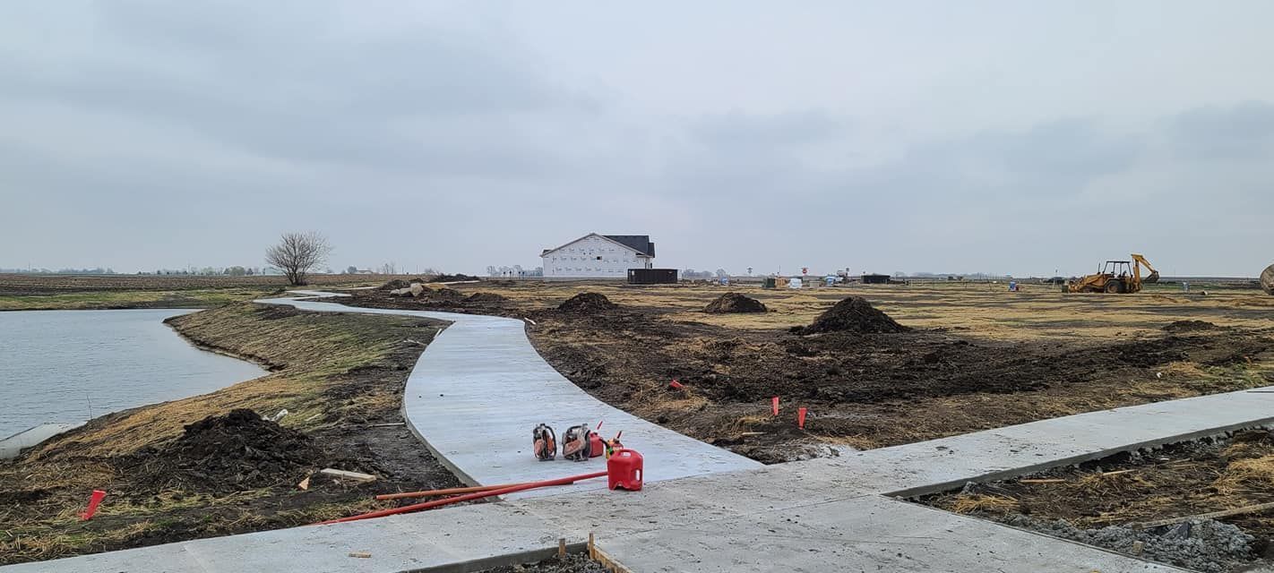 Concrete pathway curves around a pond. Construction site with piles of dirt and a building in the distance. Overcast sky.