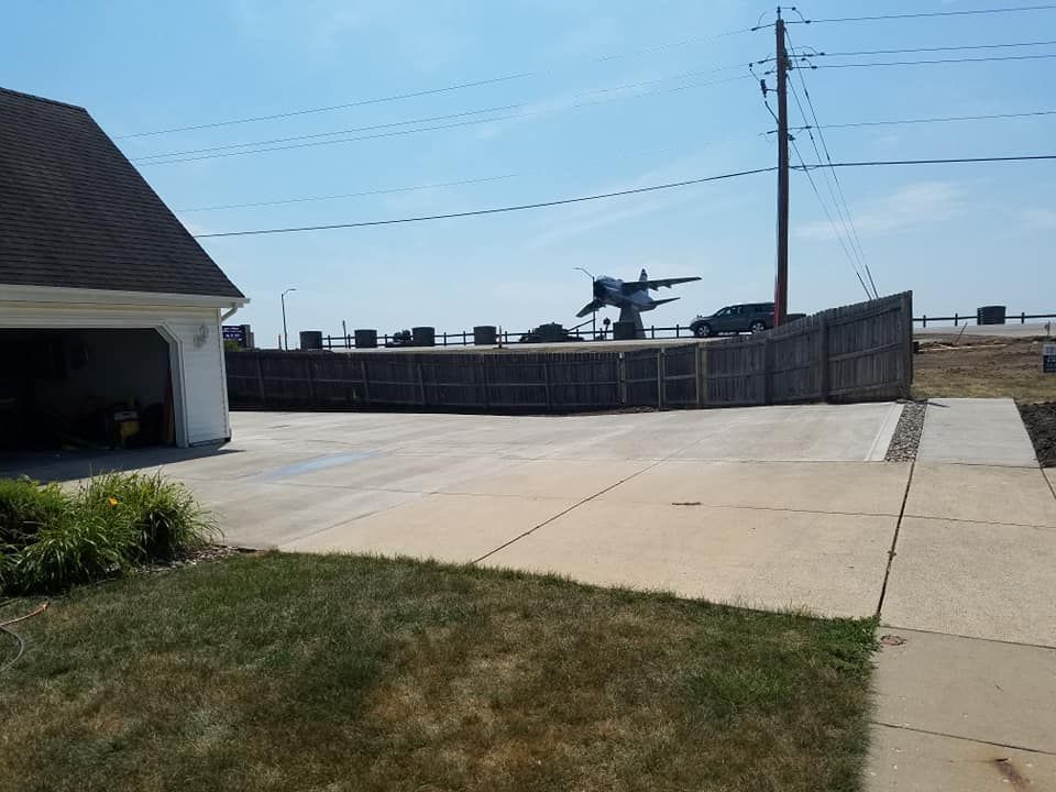 Garage and driveway with a large aircraft behind a wooden fence, under a bright blue sky.