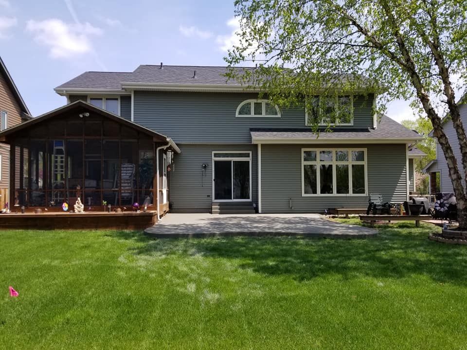 Backyard of a two-story house with a screened porch and patio. Green lawn, blue siding, and clear sky.