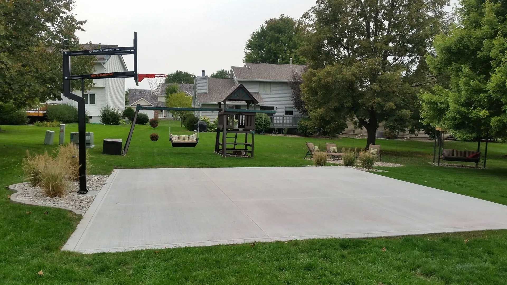 Backyard with concrete basketball court, swingset, and trees under overcast sky.