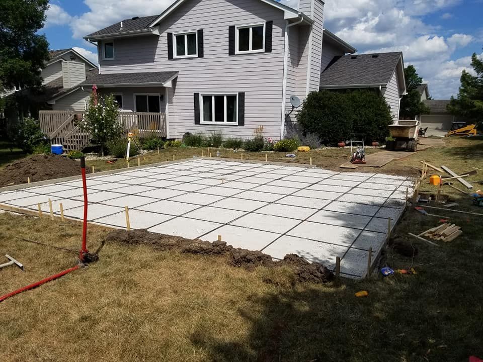 Concrete patio grid in backyard, near house. Grass, construction materials visible. Sunny day.