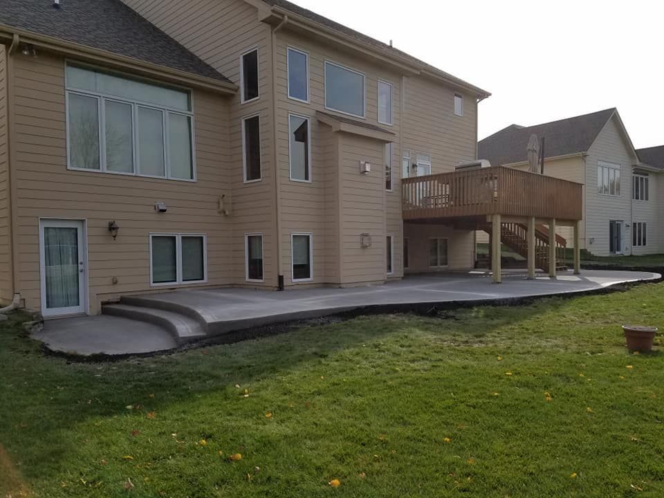 Back of a tan house with a concrete patio and a wooden deck. Green lawn surrounds the structures.
