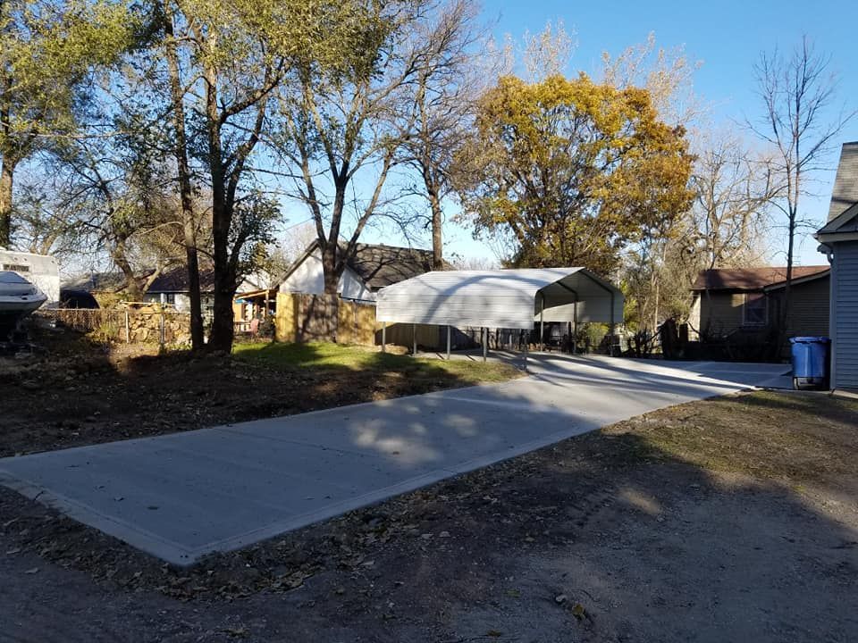 Concrete driveway leading to a metal carport under autumn trees. Blue trash can on right.