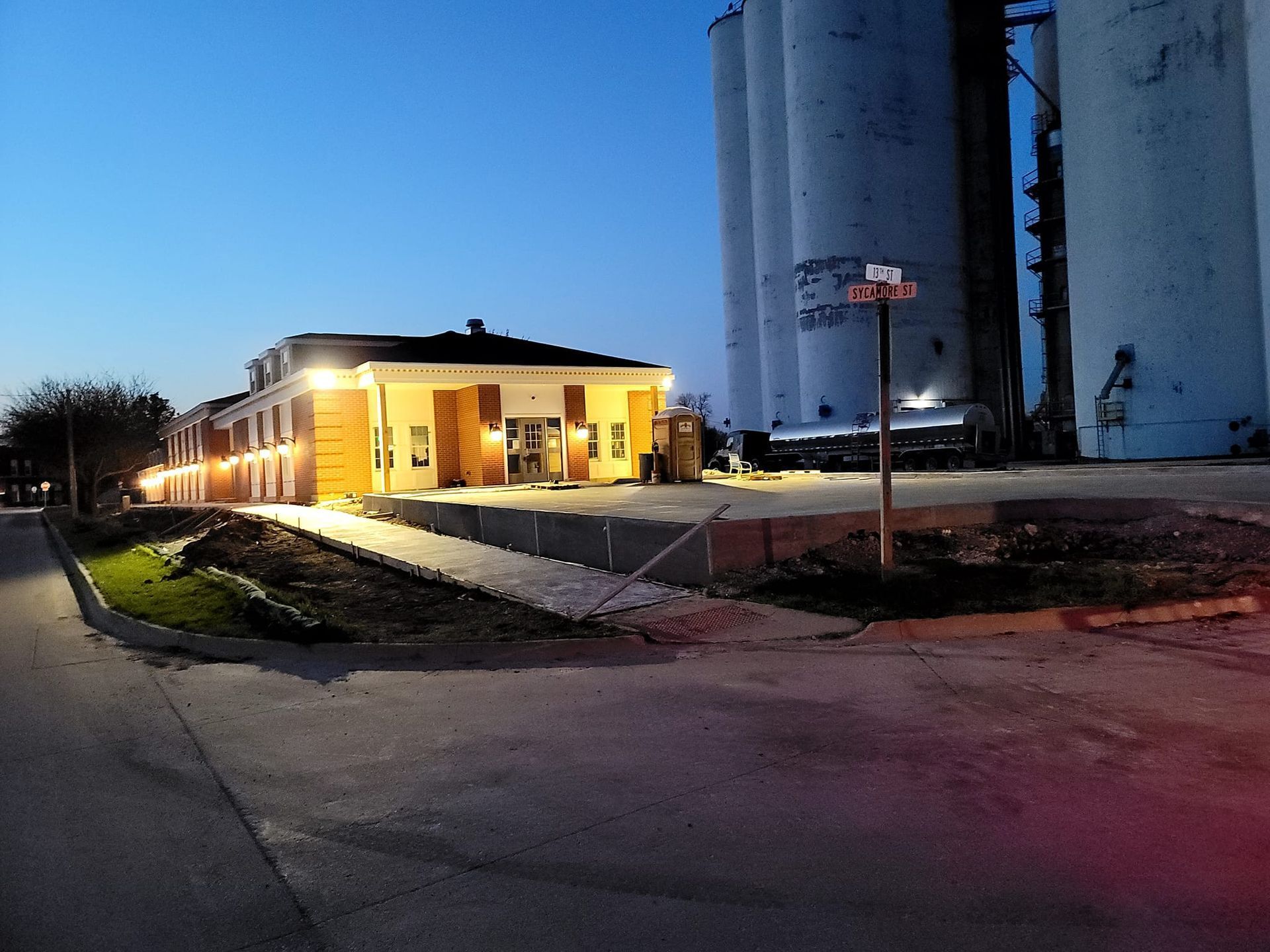 A low-rise motel at dusk next to large industrial silos. Bright lights illuminate the motel entrance and sidewalk.