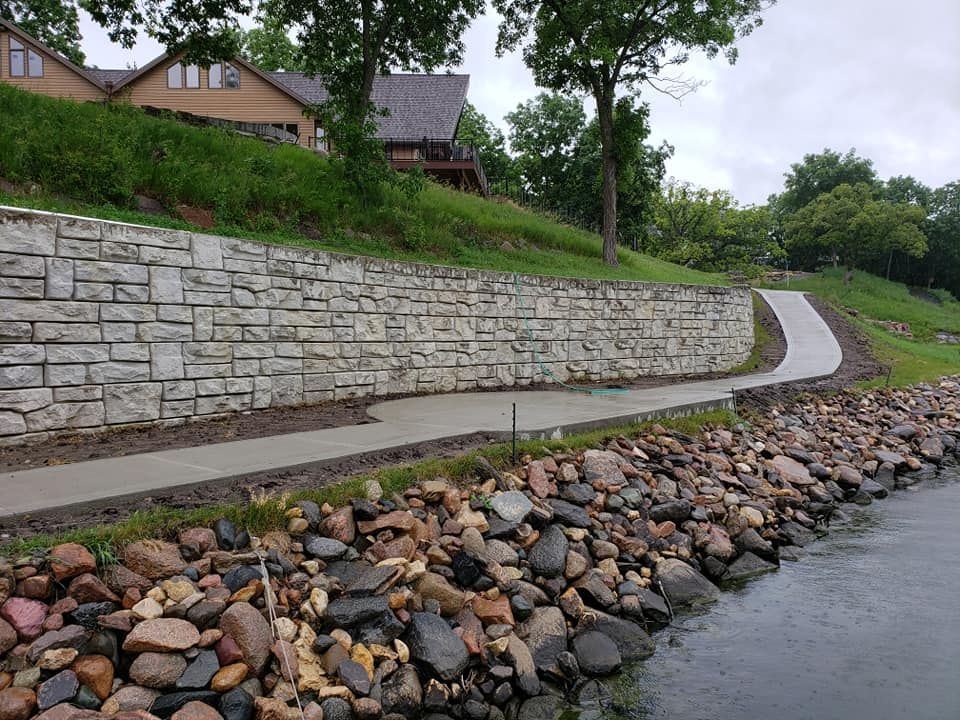 Stone retaining wall and paved path along a shoreline with rocks. Houses sit above the wall.