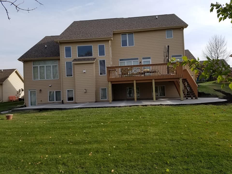 Back of two-story tan house with a wooden deck and concrete patio, set in a green yard under a cloudy sky.