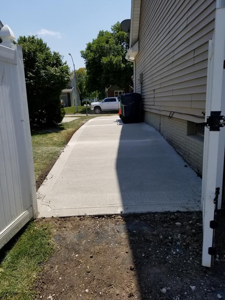 Concrete walkway next to a light-colored house. Black trash cans sit at the end of the walkway. White fence on left.