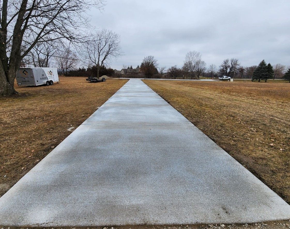 A long, straight concrete path stretches through an open, dry grass field under a cloudy, overcast sky.