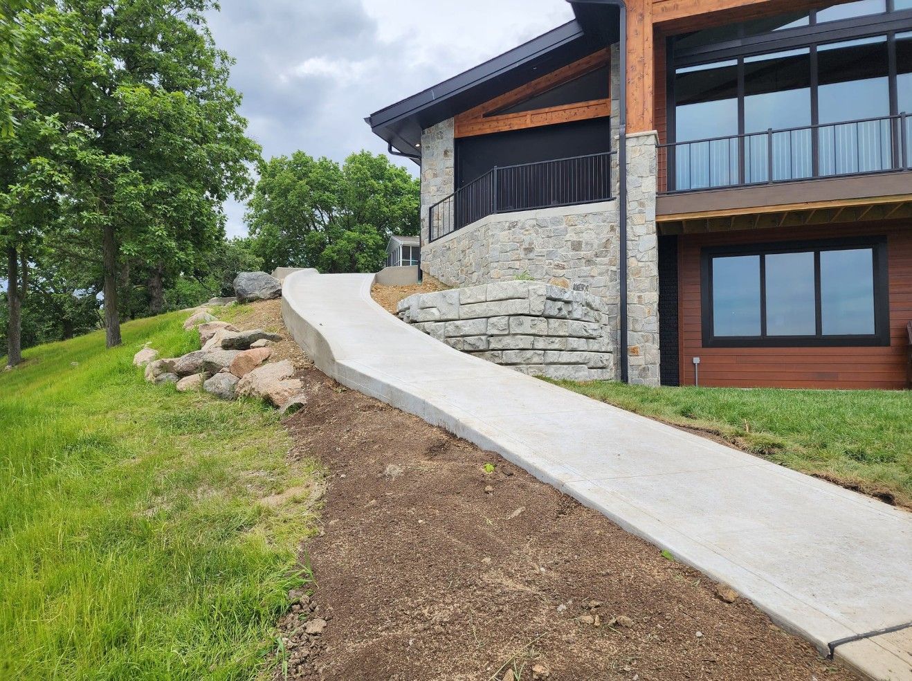 A long concrete accessibility ramp leading up to the side of a modern stone and wood house on a grassy hill.