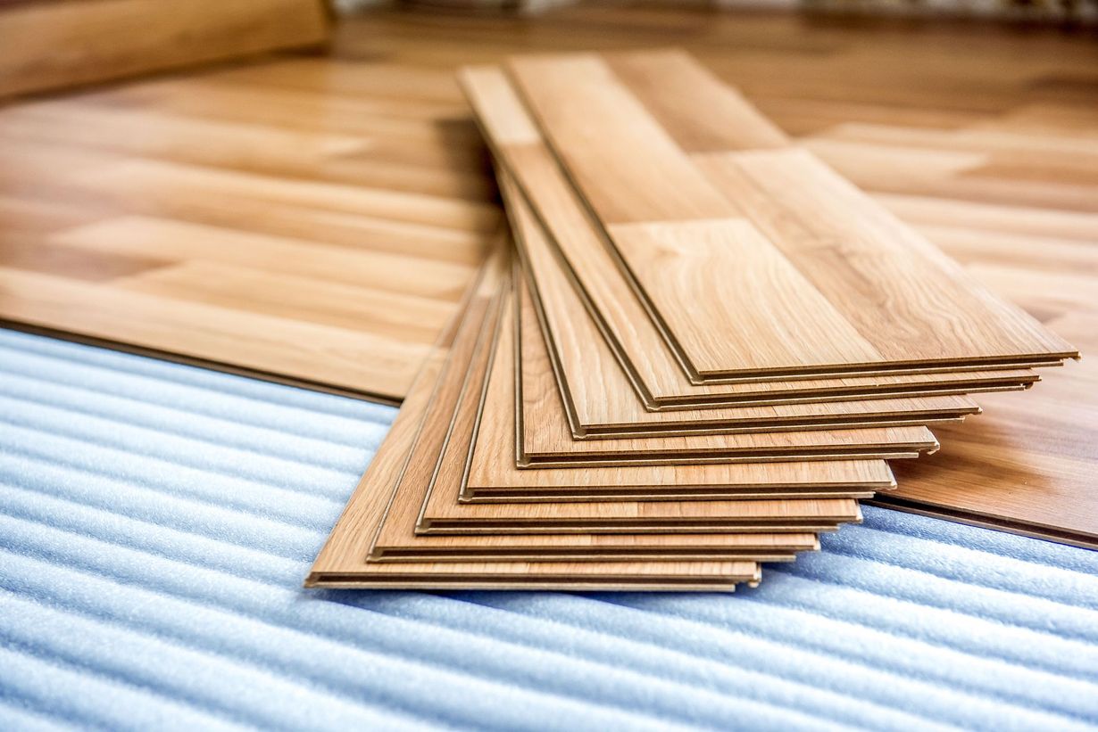 Pile of light brown laminate flooring planks on a blue-white mat, with installed flooring in the background.