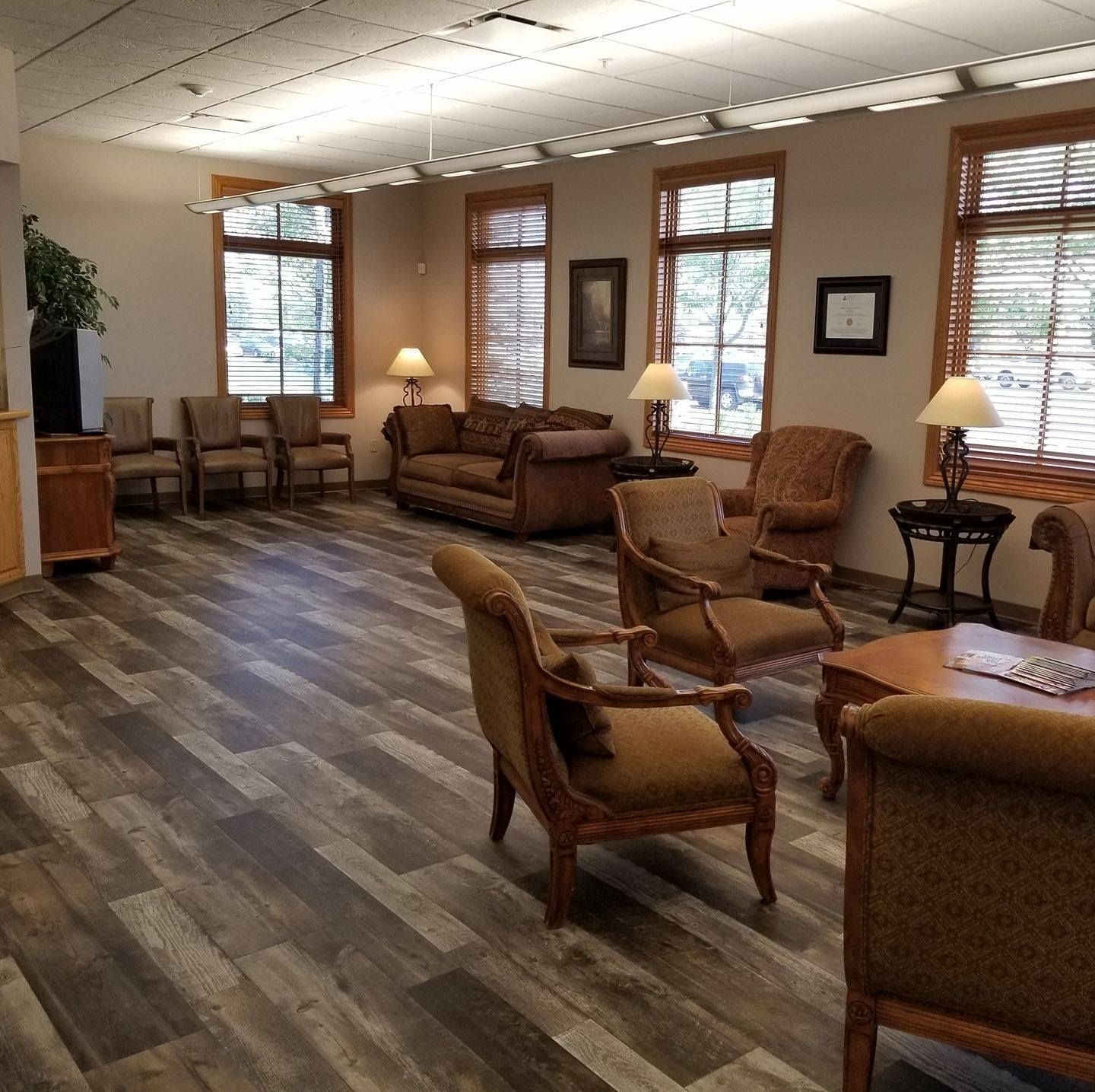 Waiting room with wood-look floor, brown furniture, windows with blinds, and table lamps.