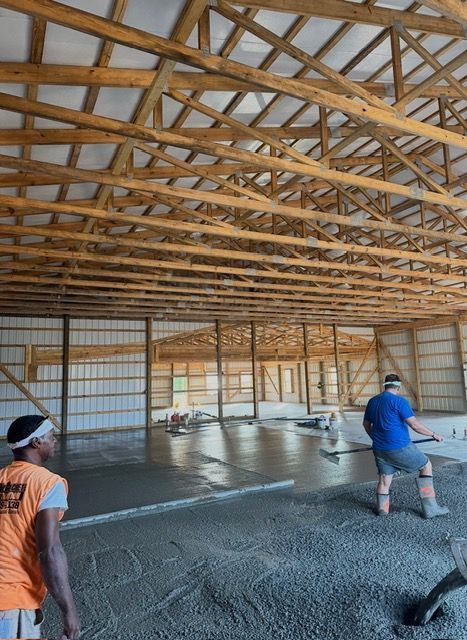 Construction workers smoothing wet concrete floor inside a large barn-like building with wooden beams.
