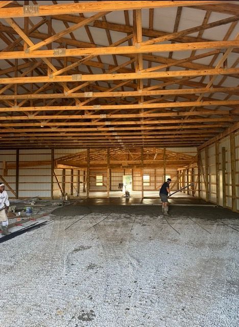 Workers spreading concrete in a large, open-air building under construction; wooden beams and trusses overhead.