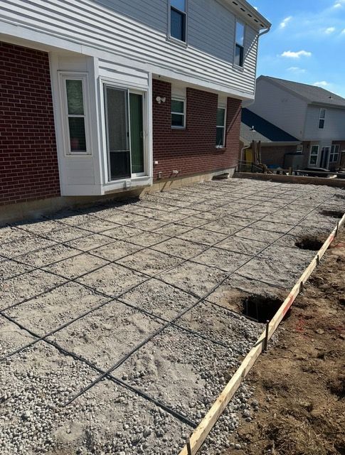 Concrete patio construction site next to a brick house. Rebar grid laid on gravel base. Wooden forms define the patio perimeter.