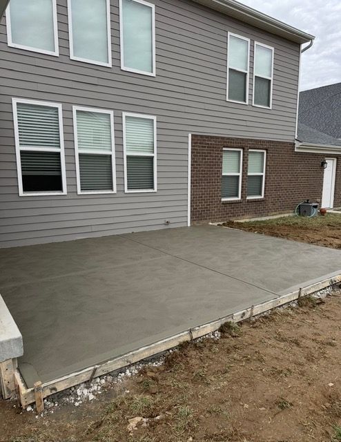 Newly poured concrete patio next to a two-story house with gray siding and brick.
