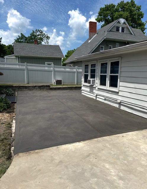 A gray concrete patio next to a light-colored house. A white fence is in the background. Blue sky with clouds.