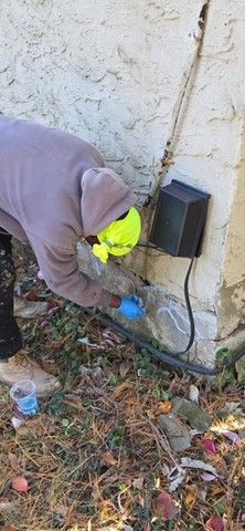 Person in gray hoodie near a building, working on a utility box, with a neon yellow item and blue gloves.
