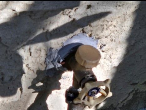 Exterior water spigot with tan handle and white wall, partially covered by grey material.