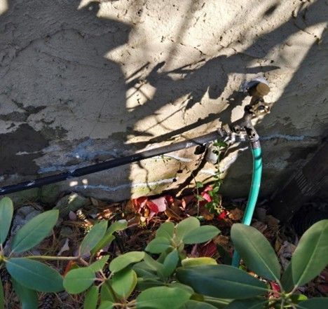 Water spigot with attached hoses, mounted on a stucco wall, with greenery in the foreground and a shadow above.